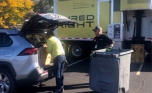 Person unloading items from the trunk of an SUV into a large bin, assisted by a worker, next to a 'SHRED RIGHT' truck, indicating a secure document destruction service.